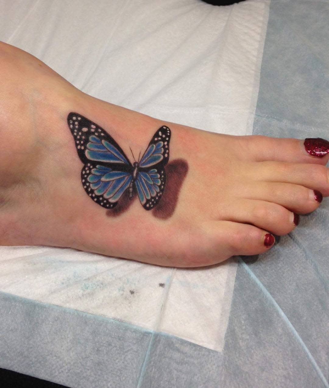 A tattoo of a blue butterfly with black and white accents on the top of a foot with red-painted toenails