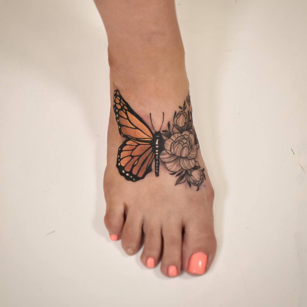 Foot tattoo of a detailed black and orange butterfly with floral designs, toenails painted in coral