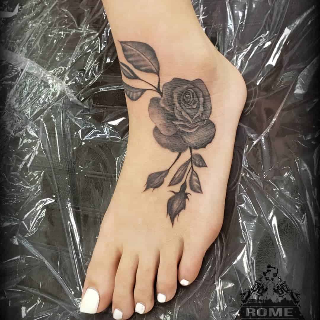 A black rose tattoo on the top of a foot with white toenails, against a transparent plastic sheet background