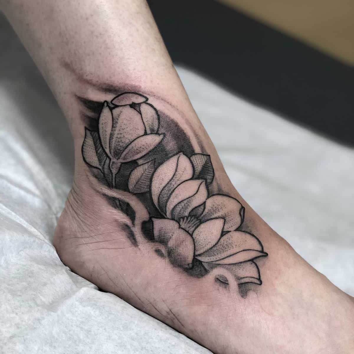 A black and grey lotus flower tattoo on an ankle with intricate shading and detail