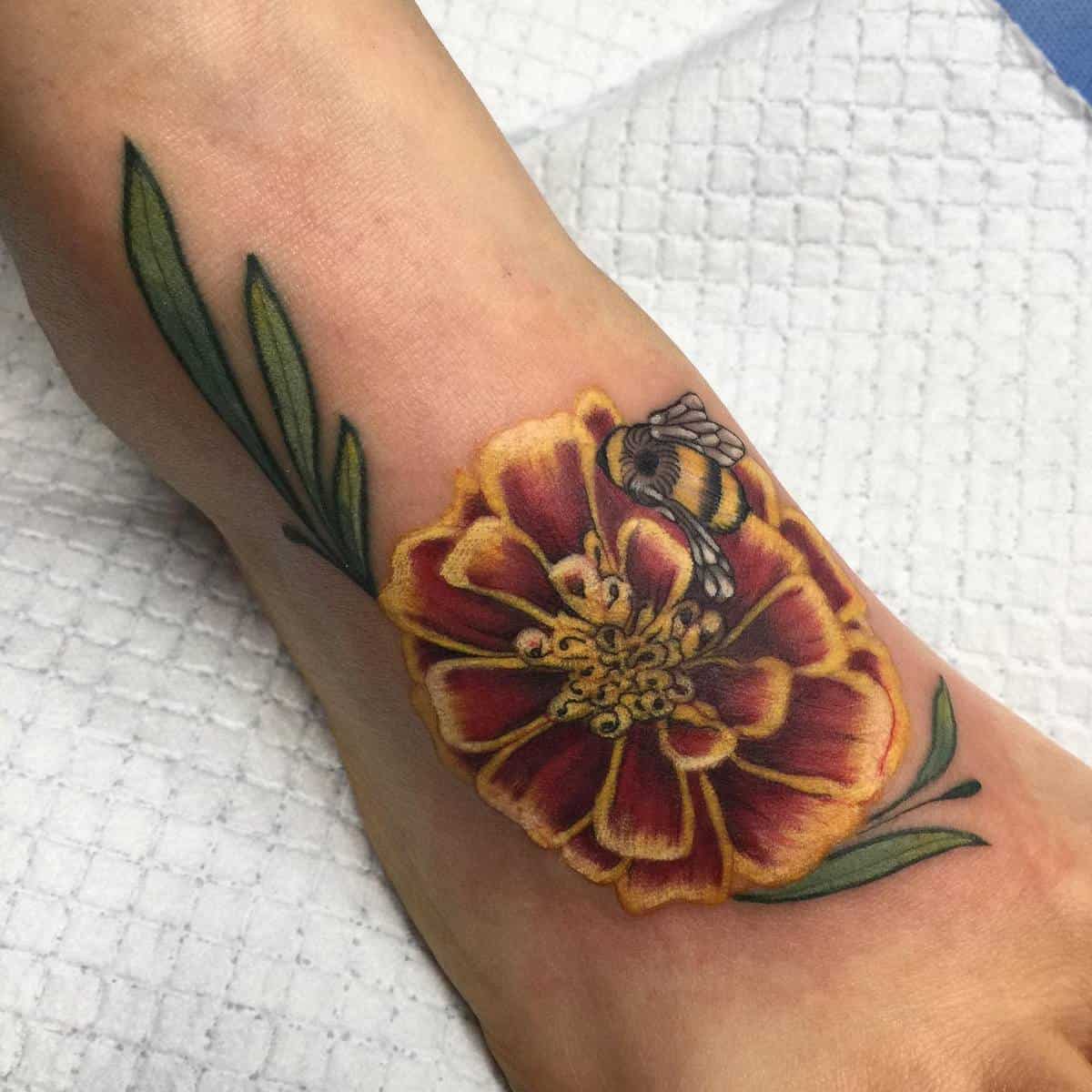 A vibrant flower tattoo with a bee on the foot, featuring green leaves and detailed yellow and red petals