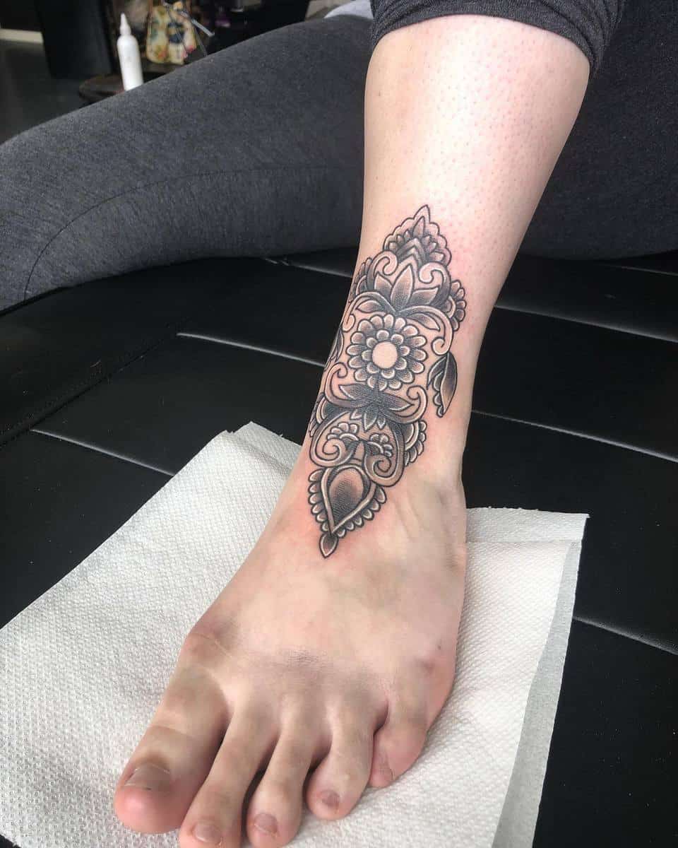 Ornate black and grey mandala tattoo on an ankle and foot, leg resting on a white paper towel