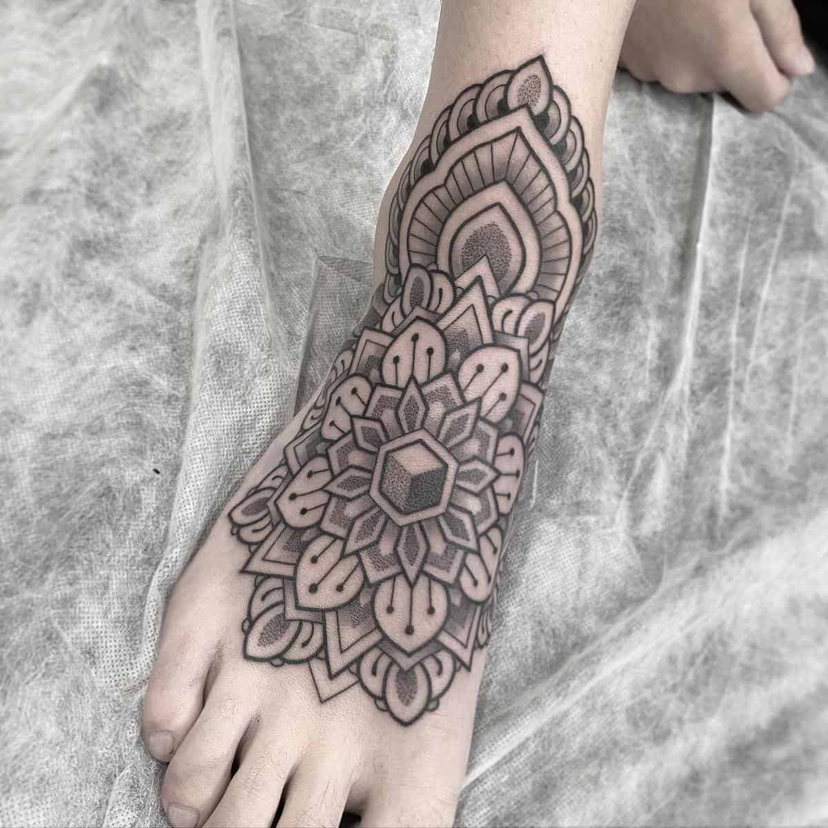 A foot with an intricate, symmetrical mandala tattoo covering the top and ankle, featuring lines and shading in black ink