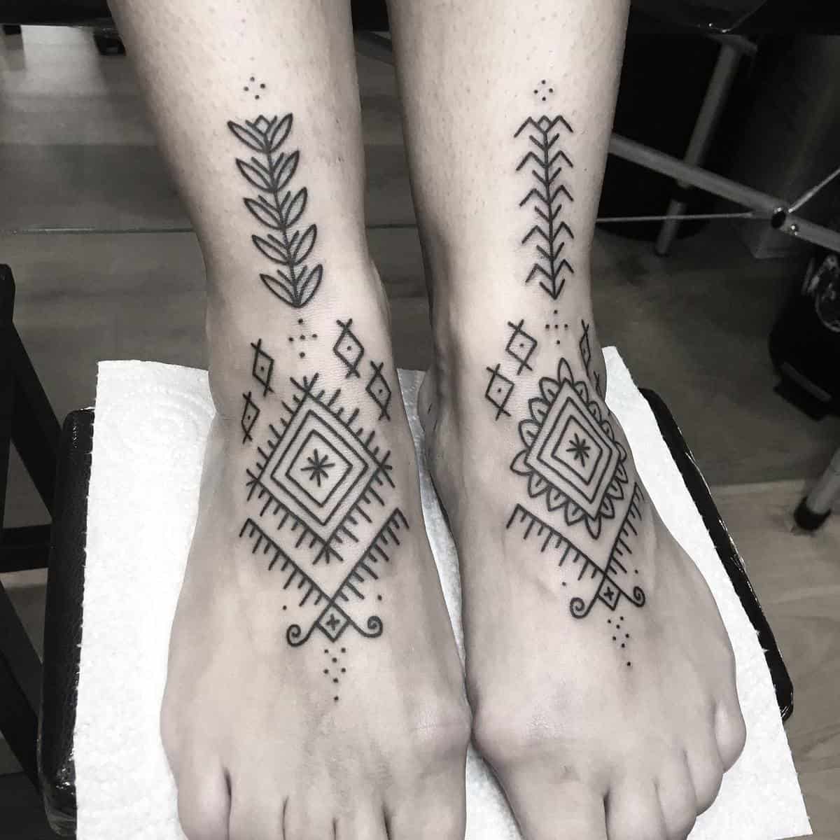 Feet with black geometric tattoos featuring diamonds, lines, and leaves placed on a white sheet in a tattoo studio
