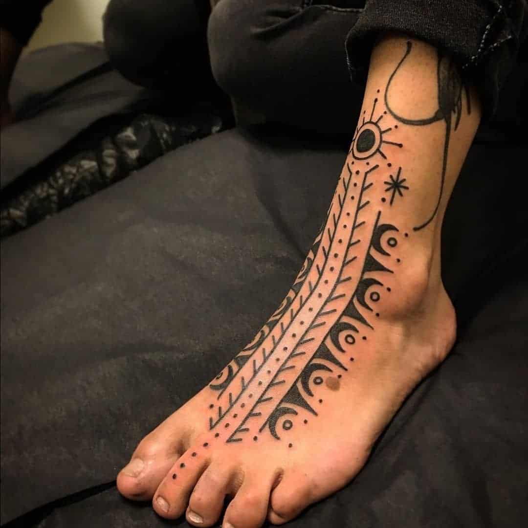 A foot with an intricate black tattoo design featuring abstract tribal patterns and symbols extending from the toes to the ankle