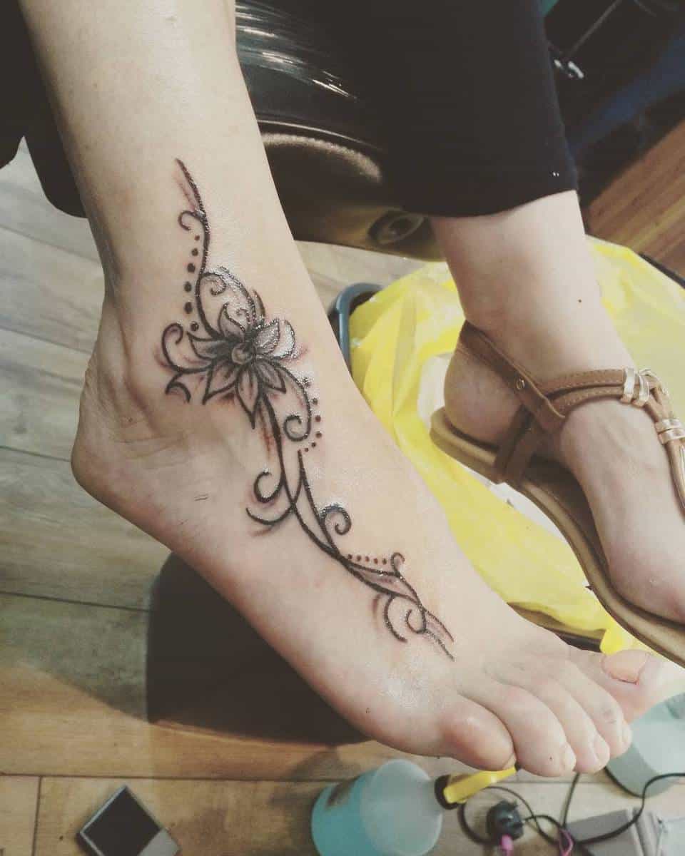 A foot tattoo featuring a floral design with elegant swirls and dots, placed on the outer part of the ankle