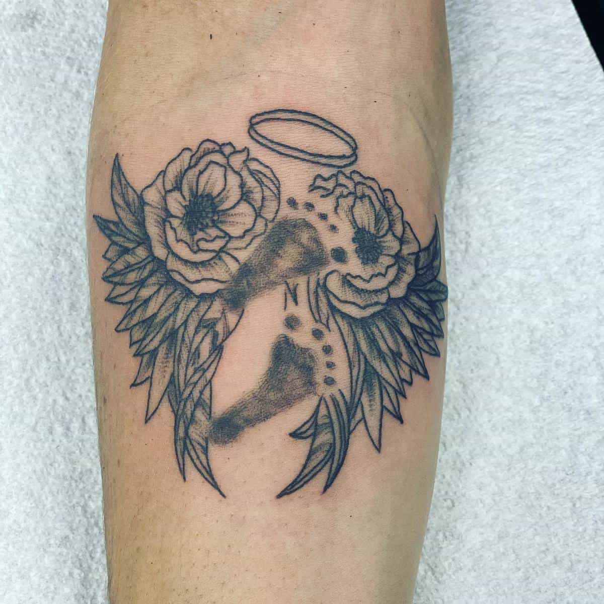 Tattoo of angel wings, flowers, a halo, and footprints on an arm representing a memorial theme