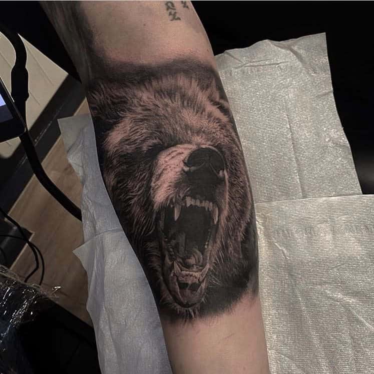 Tattoo of a majestic roaring bear head on someone's arm, displayed on a paper-covered surface