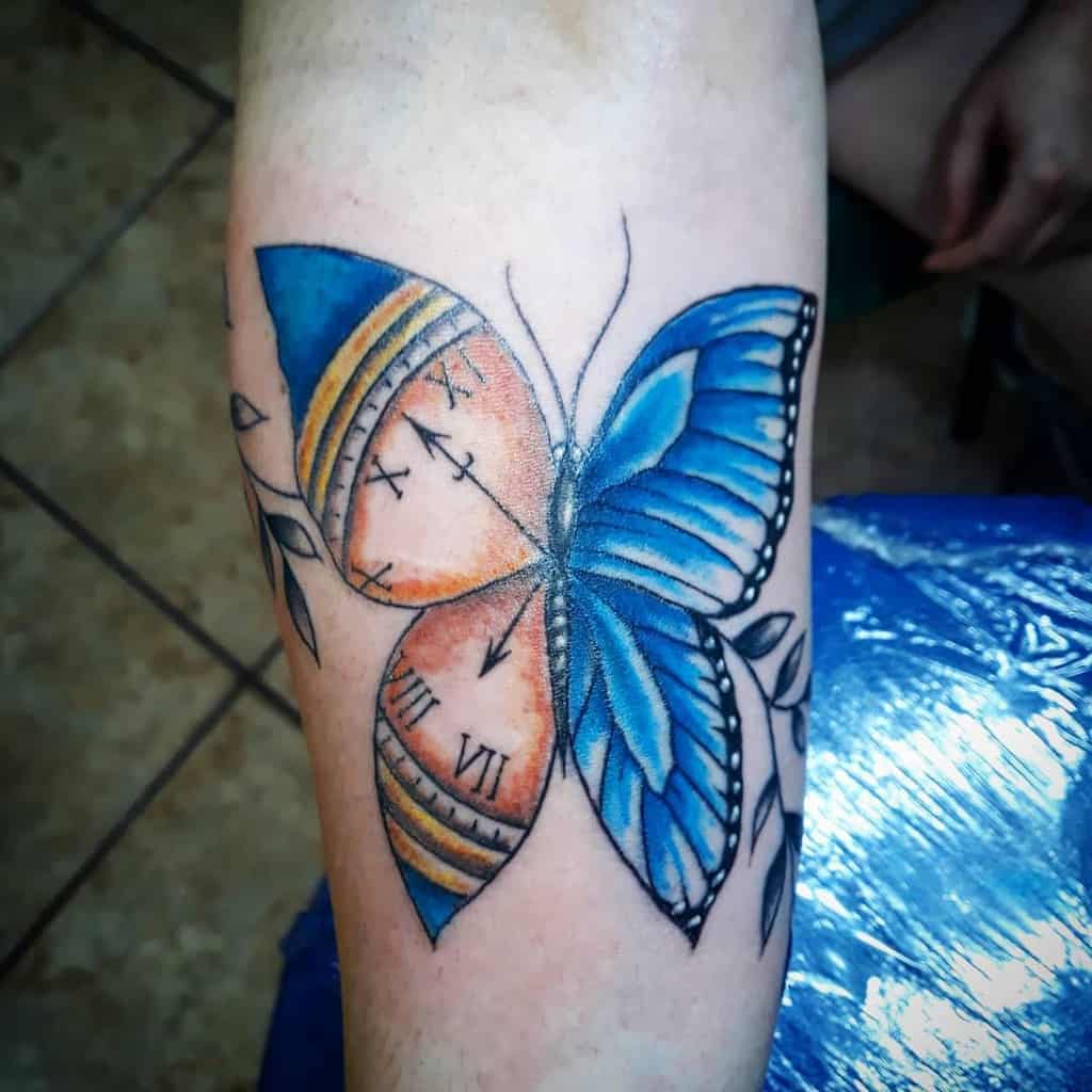 Forearm tattoo of a blue and orange butterfly with clock details on wings and leafy accents