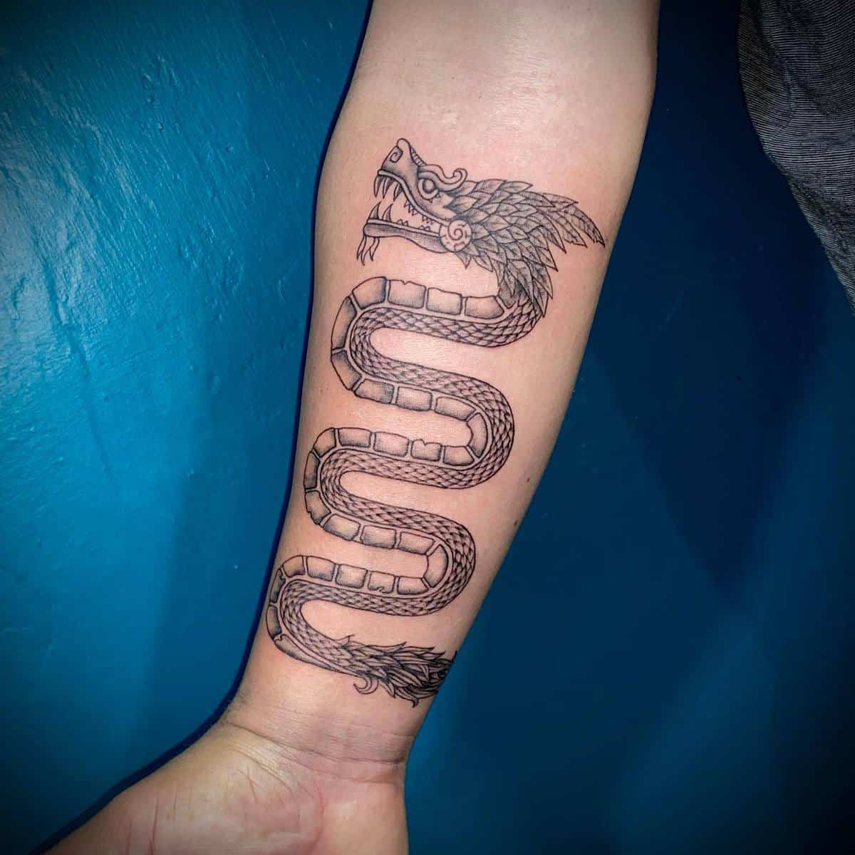 Black and gray Quetzalcoatl tattoo on the forearm featuring a coiled serpent with detailed scales and feathered head