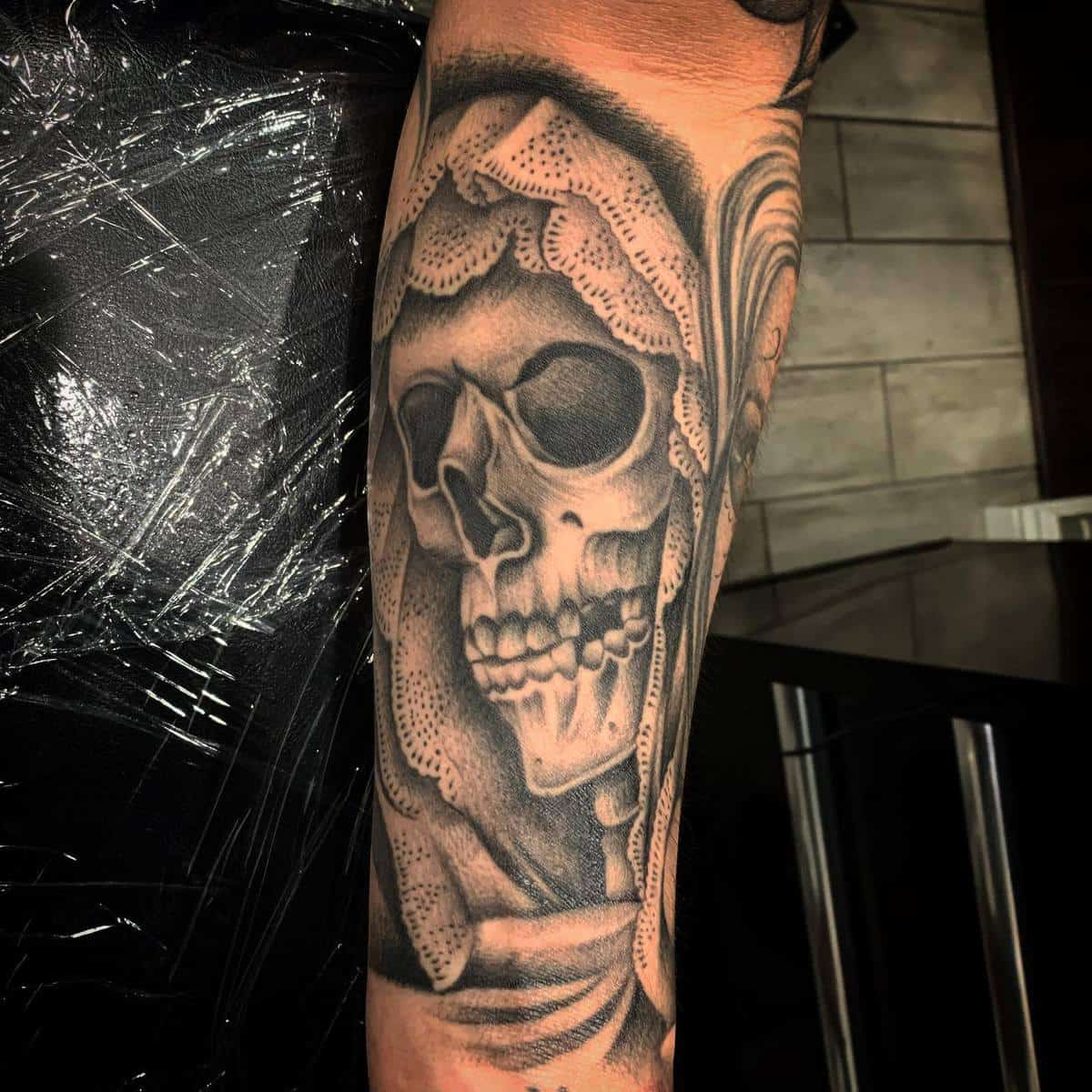 Black-and-grey tattoo of Santa Muerte on the forearm, with intricate skull details and a lace hood