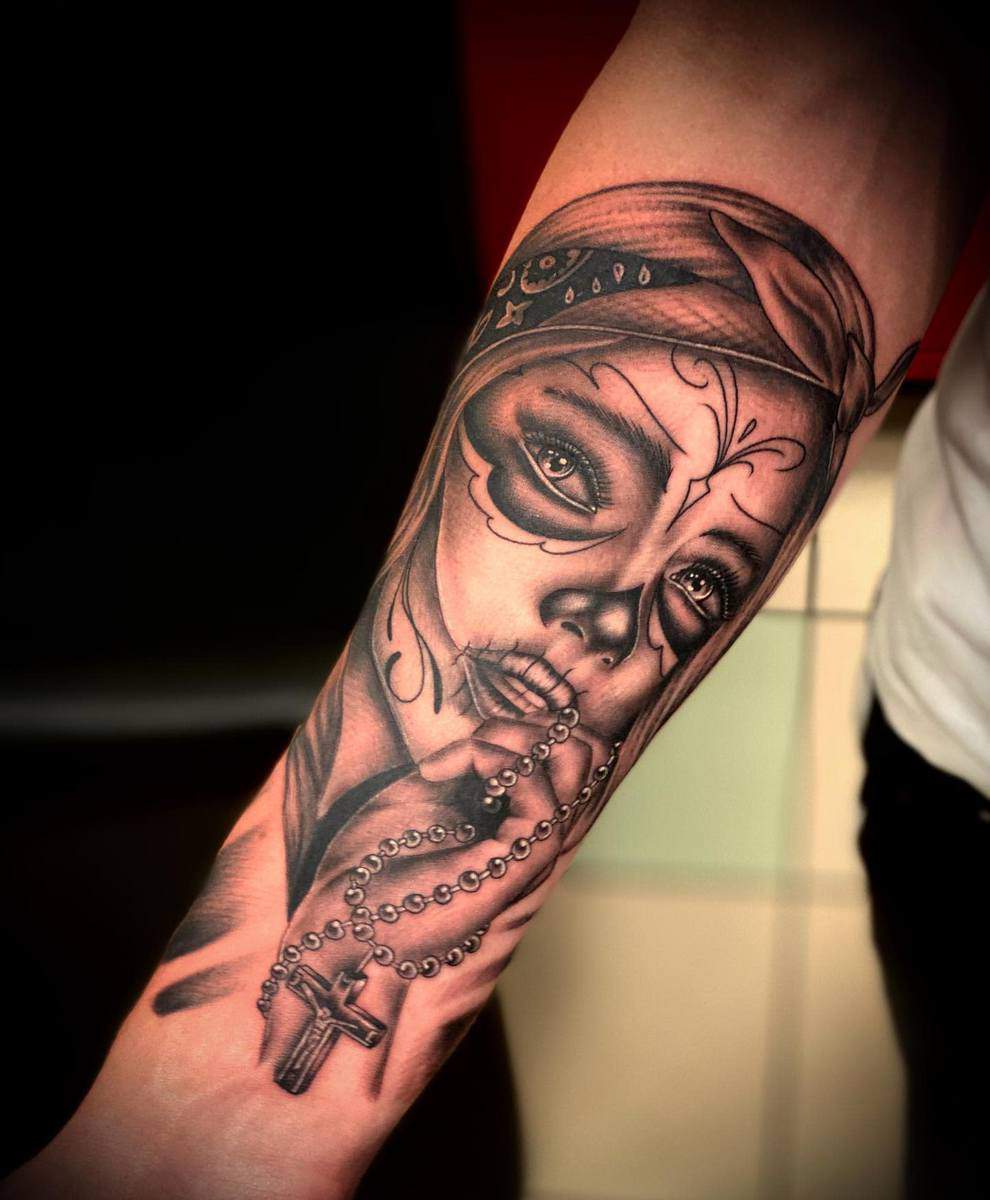 Realistic tattoo on the forearm showing a woman with Catrina makeup, clutching a rosary, blending elements of devotion and the Day of the Dead