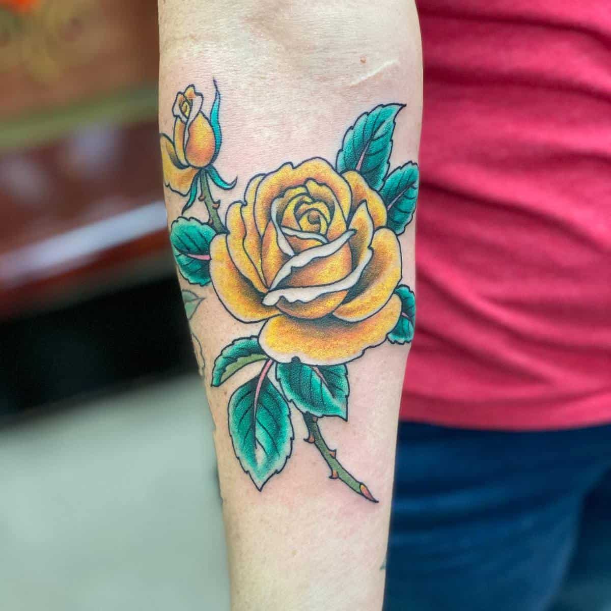 A forearm tattoo of a vibrant yellow rose with green leaves, accompanied by a rosebud