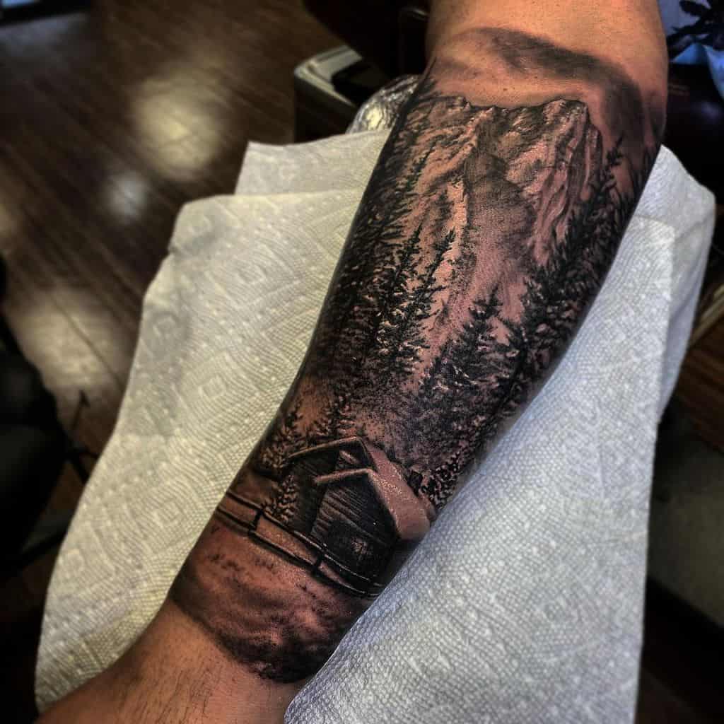 Forearm tattoo of a cabin and forest under a rugged mountain landscape