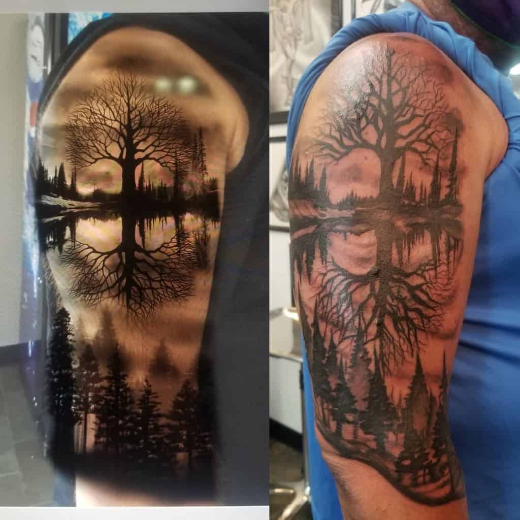 Two black ink arm tattoos showcasing a large tree with its reflection in a lake, surrounded by pine trees in a forest