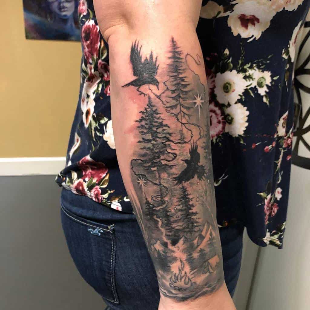 Forearm tattoo featuring crows, trees, and stars with a campfire at the bottom