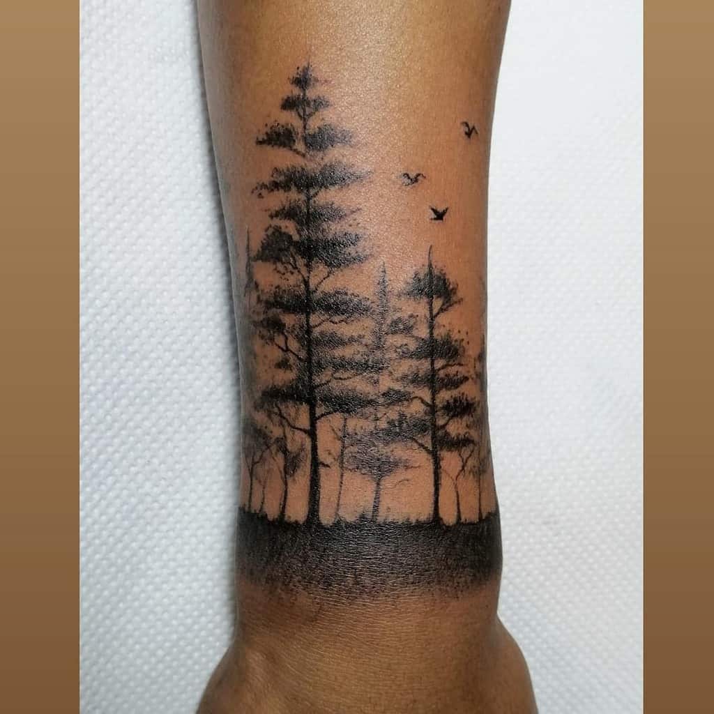 Forearm tattoo with trees, birds, and a forest silhouette in black ink