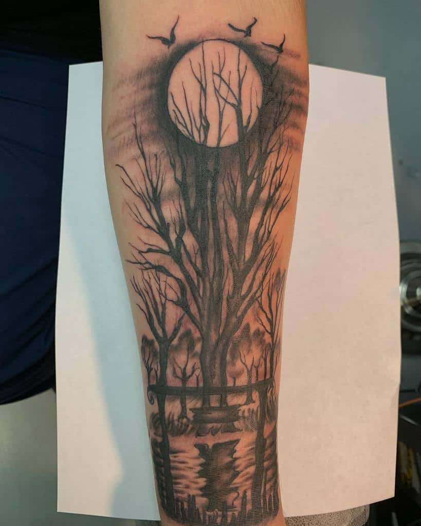 Forearm tattoo of a tree silhouette and bench under a full moon, with birds flying overhead