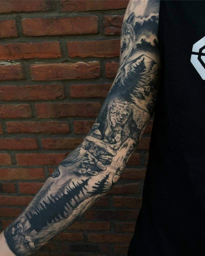 Black-and-grey full-sleeve tattoo showcasing a wolf, a lynx, and a dense pine forest under a dramatic cloudy sky, representing wilderness and strength