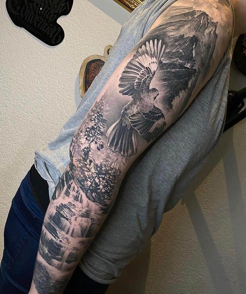 Intricate black-and-grey full-sleeve tattoo depicting a soaring bird in flight, detailed forest landscapes, cascading waterfalls, and towering mountains in the background