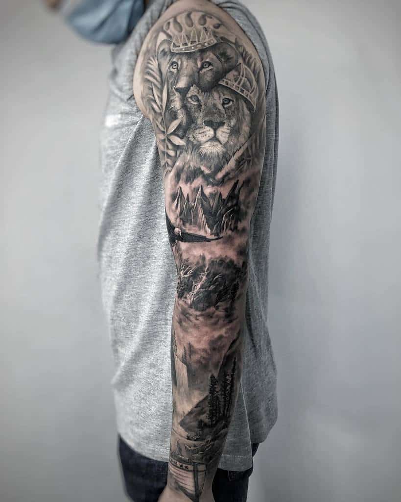 Black-and-grey full-sleeve tattoo featuring two crowned lions surrounded by laurel leaves, with a detailed landscape below showcasing a serene forest, rugged mountains, and misty clouds
