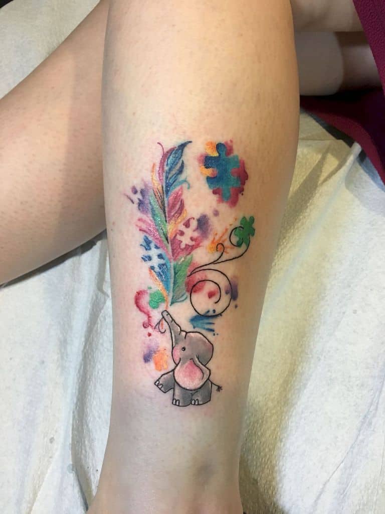 : Full color calf tattoo with baby elephant holding watercolor feather with puzzle pieces and line work flourishes.