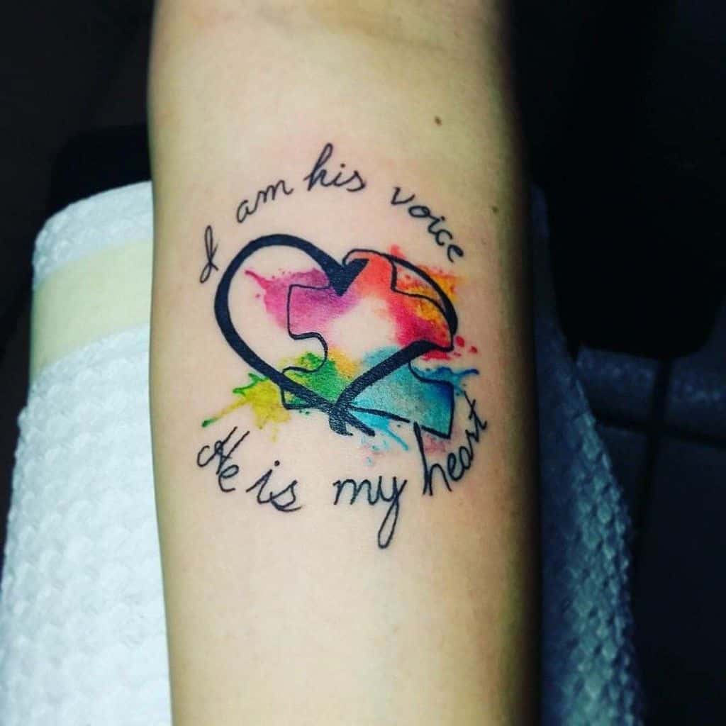 Full color forearm tattoo of a puzzle piece, and heart with watercolor splashes and an inspirational quote.