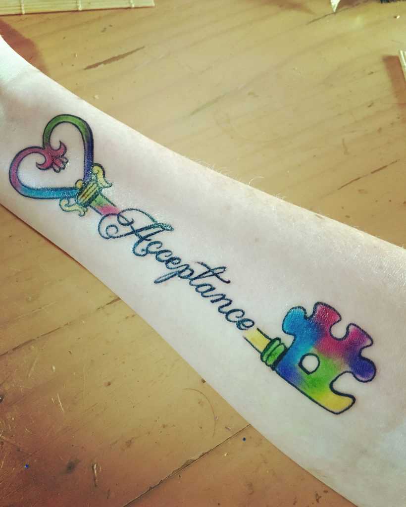 Full color forearm tattoo of a multi-colored puzzle key with text: Acceptance.