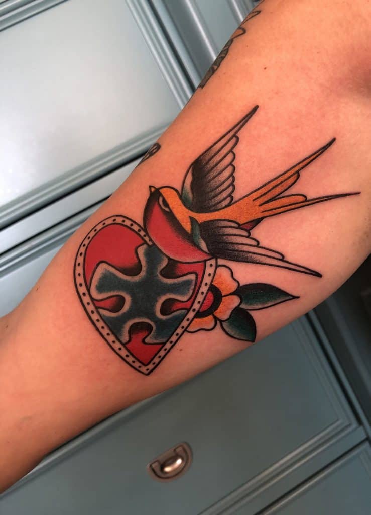Full color forearm tattoo of a puzzle piece inside a heart with an American traditional swallow and flower.