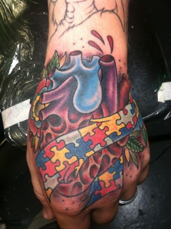 Full color hand tattoo of anatomical hear wrapped in multi-colored puzzle piece scrolls.