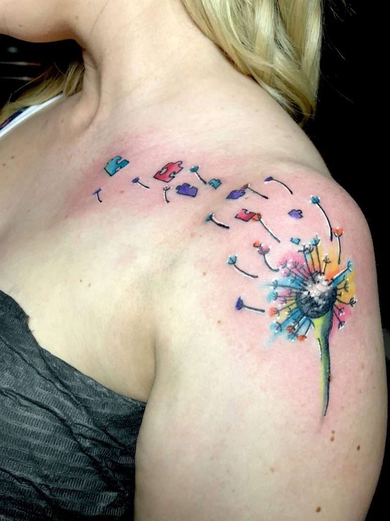 Full color upper-arm tattoo of a multi-colored dandelion with seeds and puzzle pieces blowing in the wind.