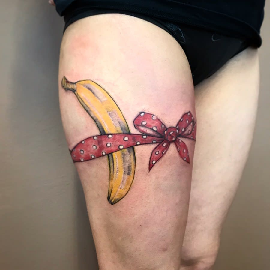 Tattoo of a banana wrapped in a red bow with white polka dots on an upper leg