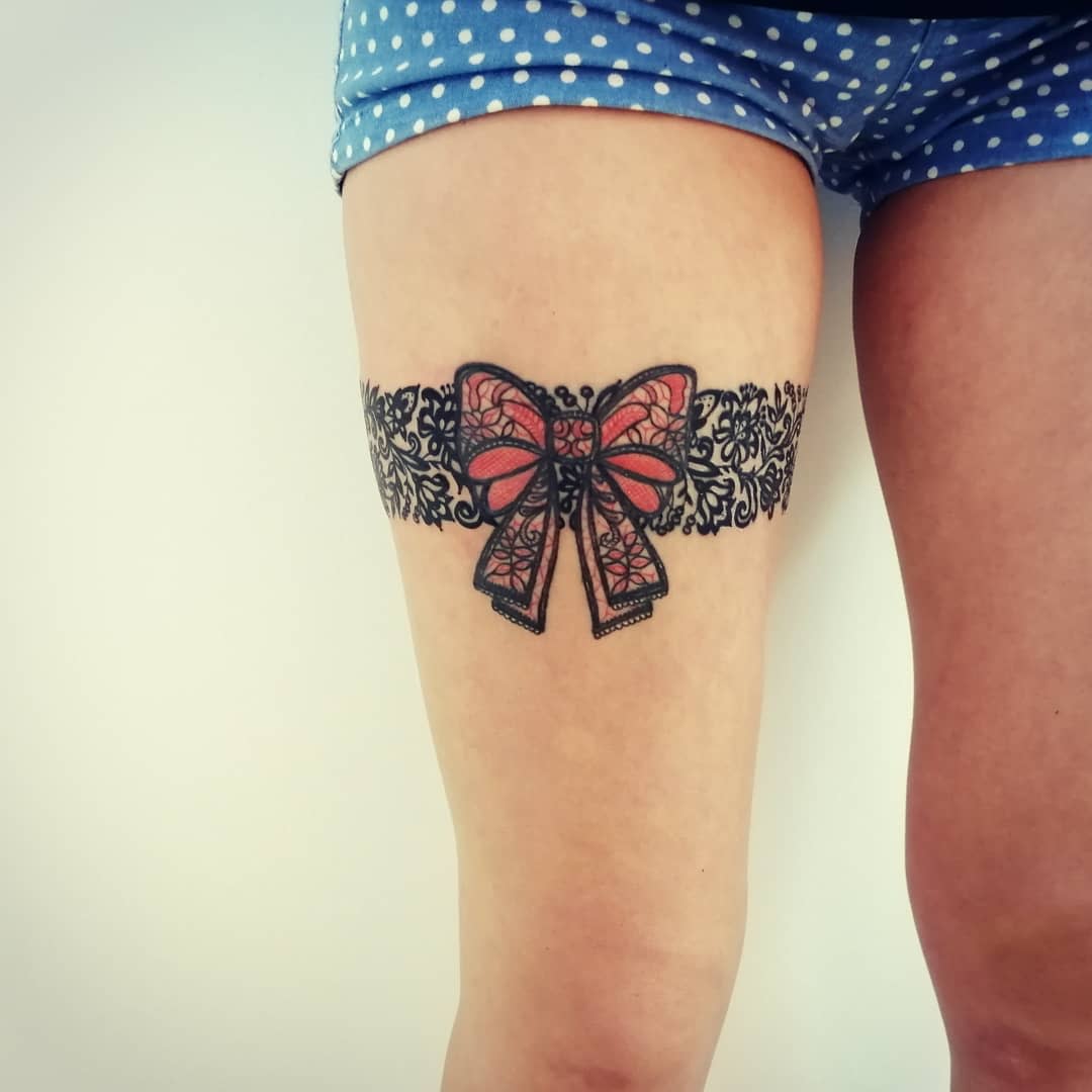 A leg tattoo of a detailed lace garter with a red bow on the thigh