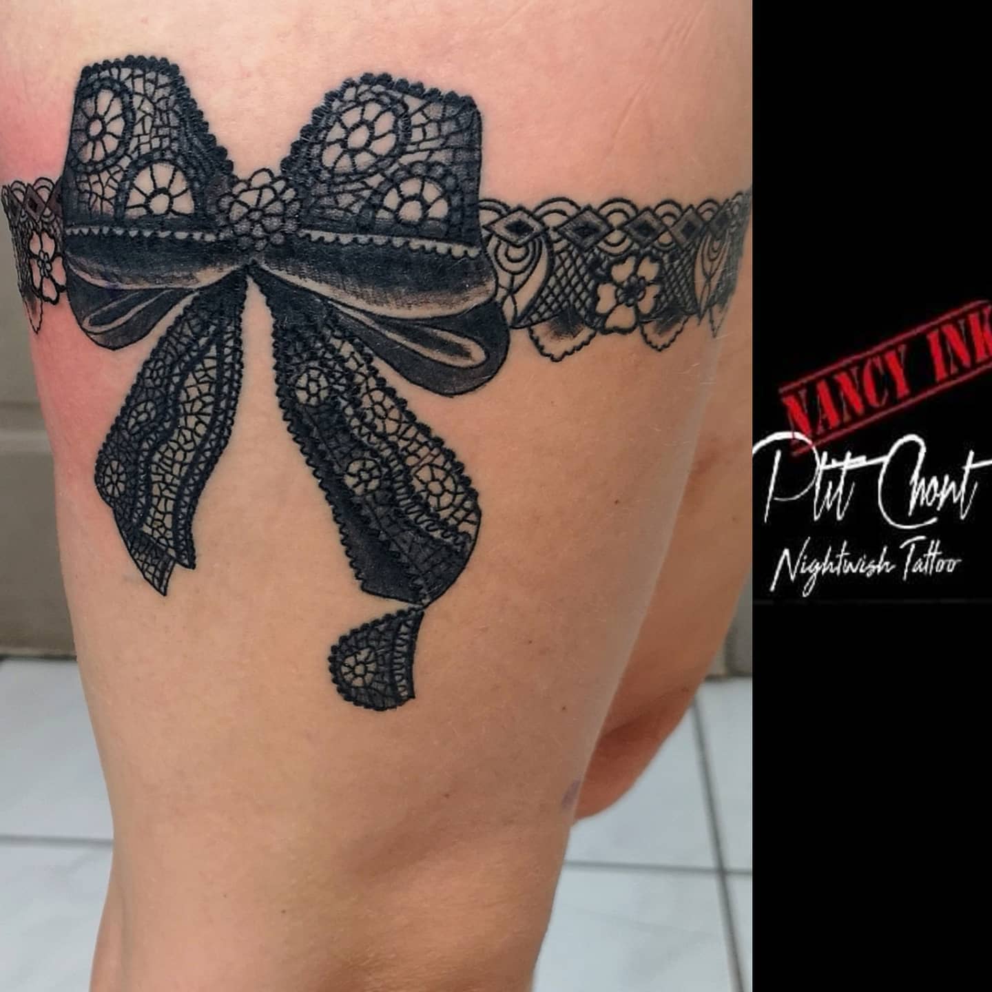 Tattoo of a black lace bow encircling a thigh, with intricate lace patterns and detailing
