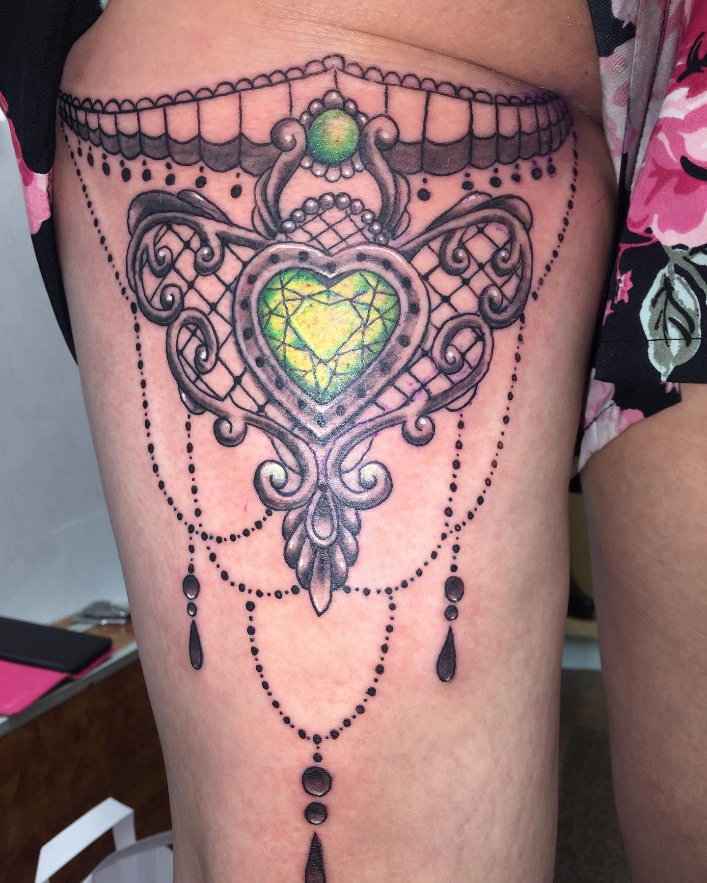 Intricate thigh tattoo with a green gem centerpiece and ornate, symmetrical designs in black ink