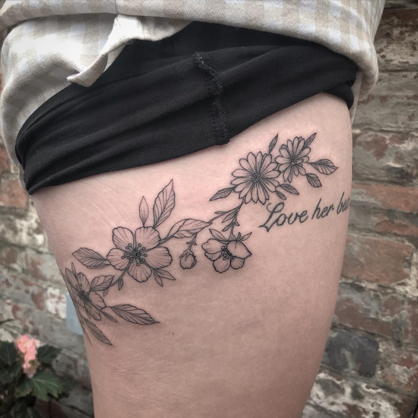 Black line floral tattoo with "Love her but" text on an upper thigh, near brick wall