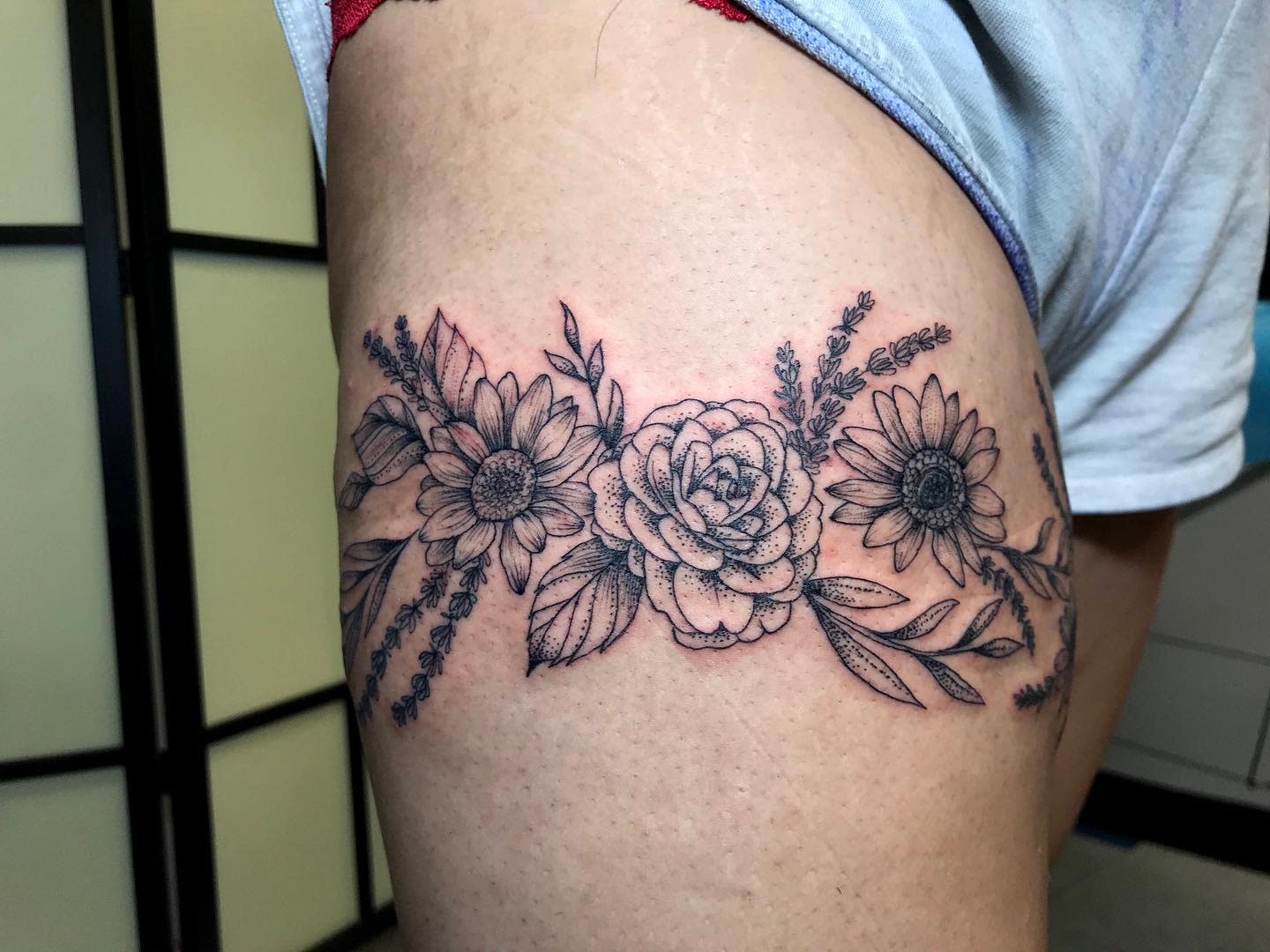 Black and grey tattoo of flowers, including sunflowers and roses, on an upper thigh