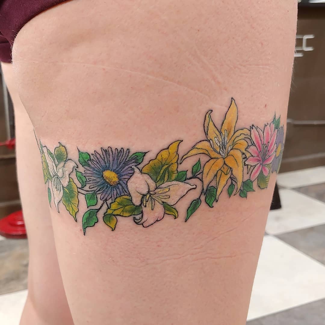 A thigh tattoo featuring colorful flowers including lilies, daisies, and leaves in a band design