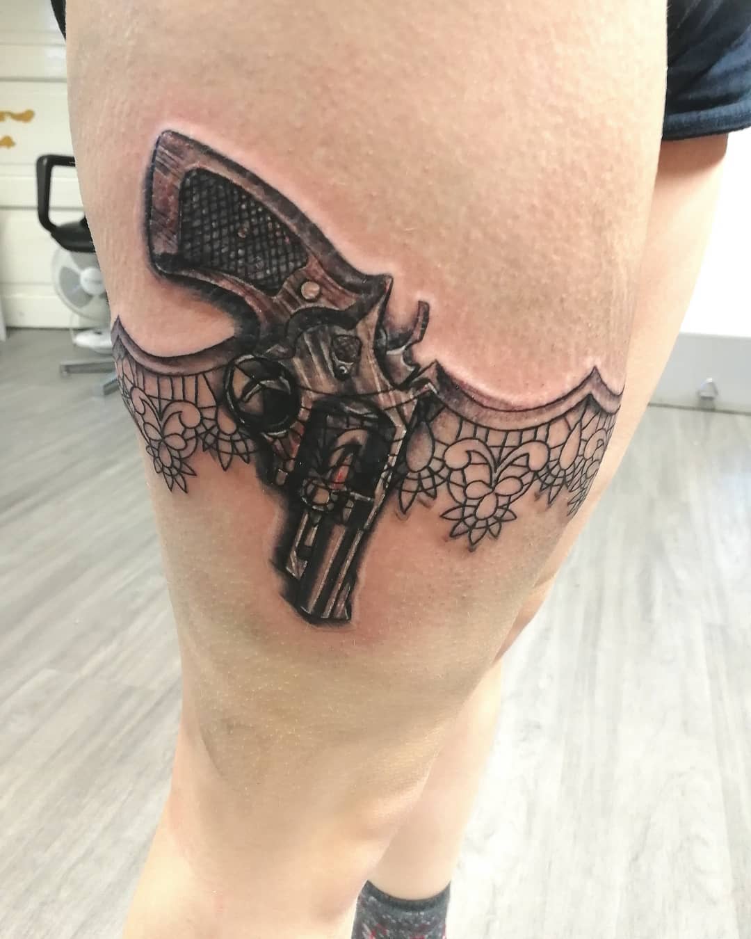 A realistic revolver tattoo with lace garter detail on a thigh