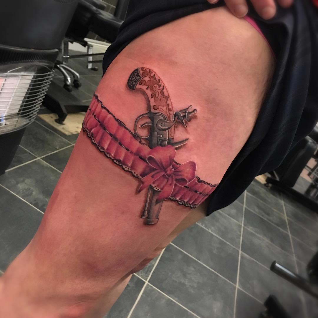 Tattoo of a gun tucked into a pink garter with a bow on an upper thigh