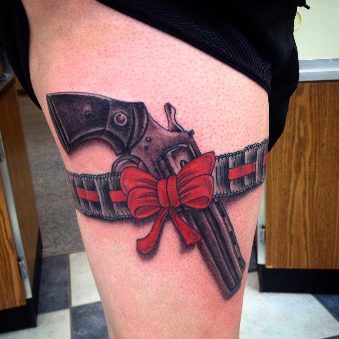 Tattoo of a revolver tucked in a garter belt with a red ribbon bow on the upper thigh