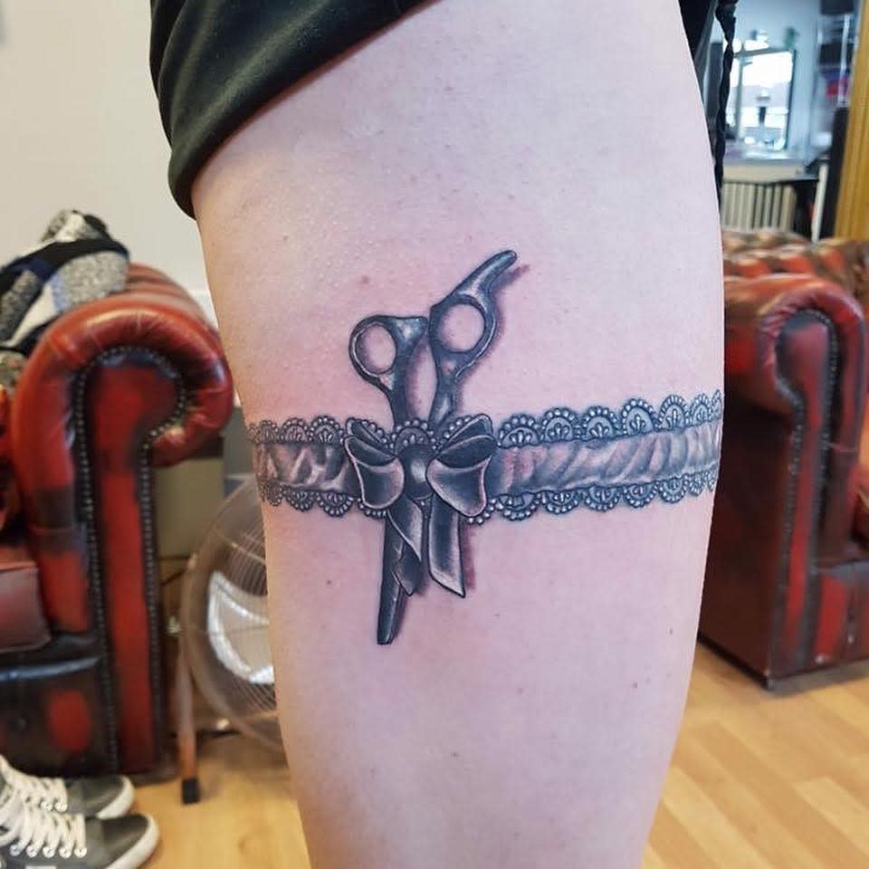 Tattoo of lace garter with bow and scissors on the side of a thigh