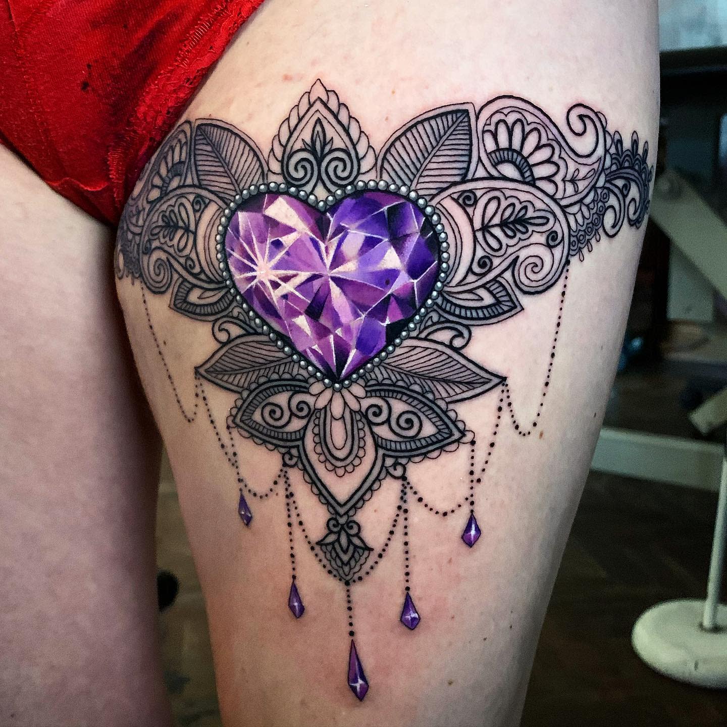 Ornate tattoo with a faceted purple heart and intricate lacework design on upper thigh