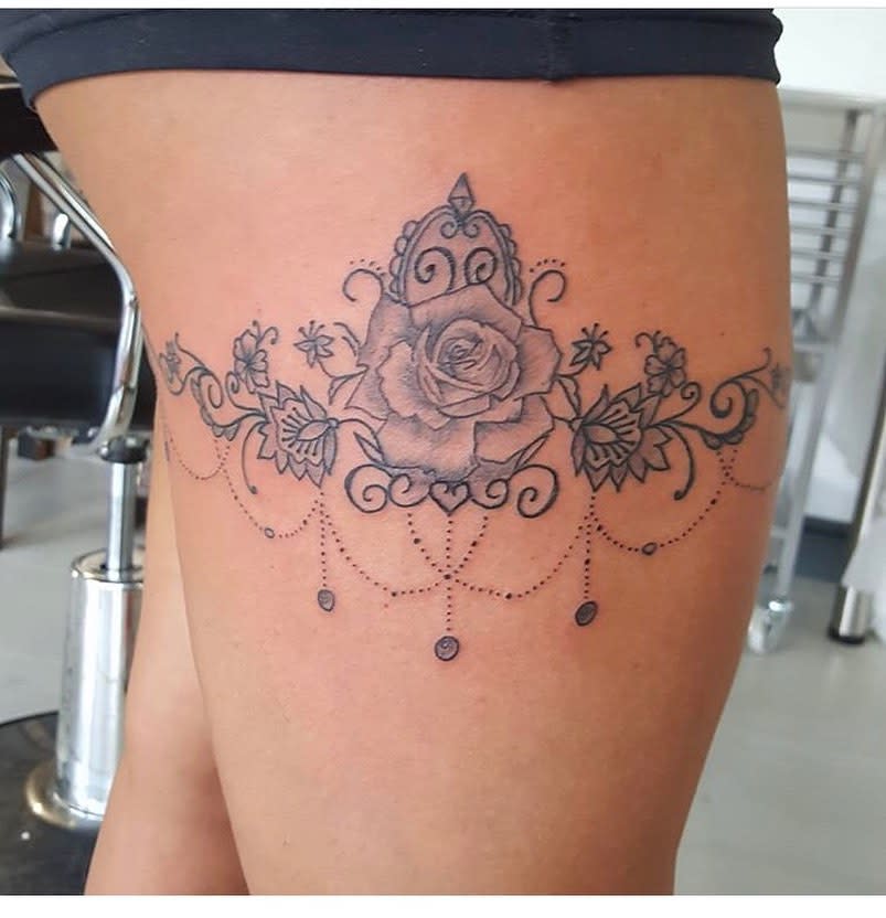 Intricate thigh tattoo featuring a central rose, ornate swirls, and hanging beads or jewels