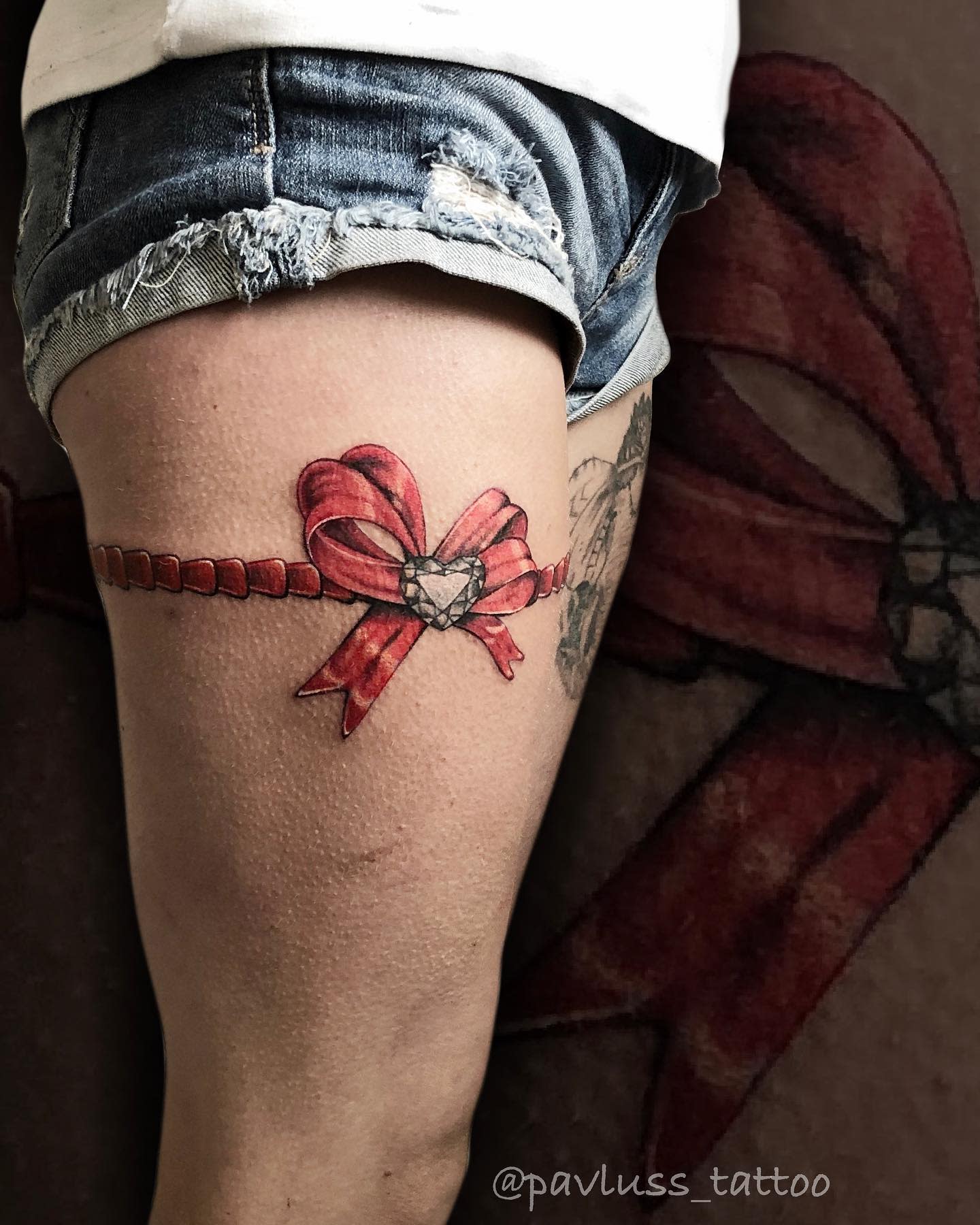 Close-up of a thigh tattoo with a red bow and a heart-shaped gem