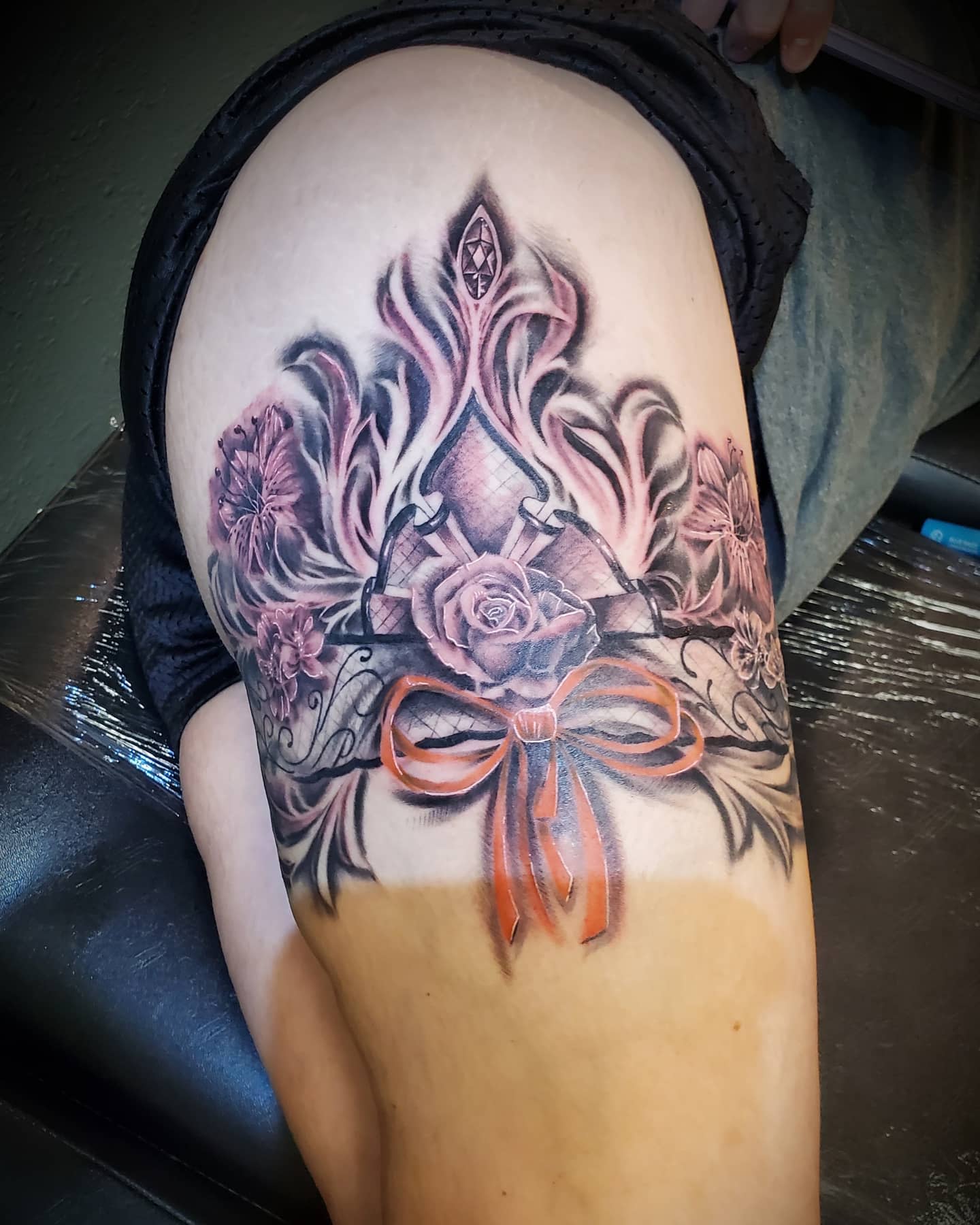 Colorful tattoo on thigh featuring a red ribbon, roses, and intricate floral patterns in grayscale