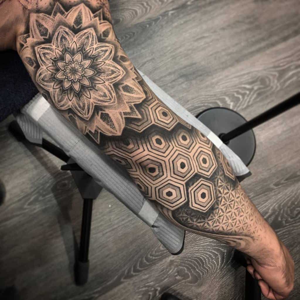 A black and grey geometric sleeve tattoo featuring a large mandala on the shoulder, hexagonal patterns on the forearm, and intricate linework