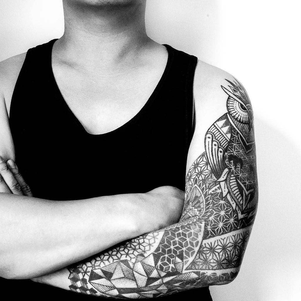 A black and grey geometric half sleeve tattoo featuring detailed mandala designs, intricate line patterns, and hexagonal shapes, wrapping around the upper arm