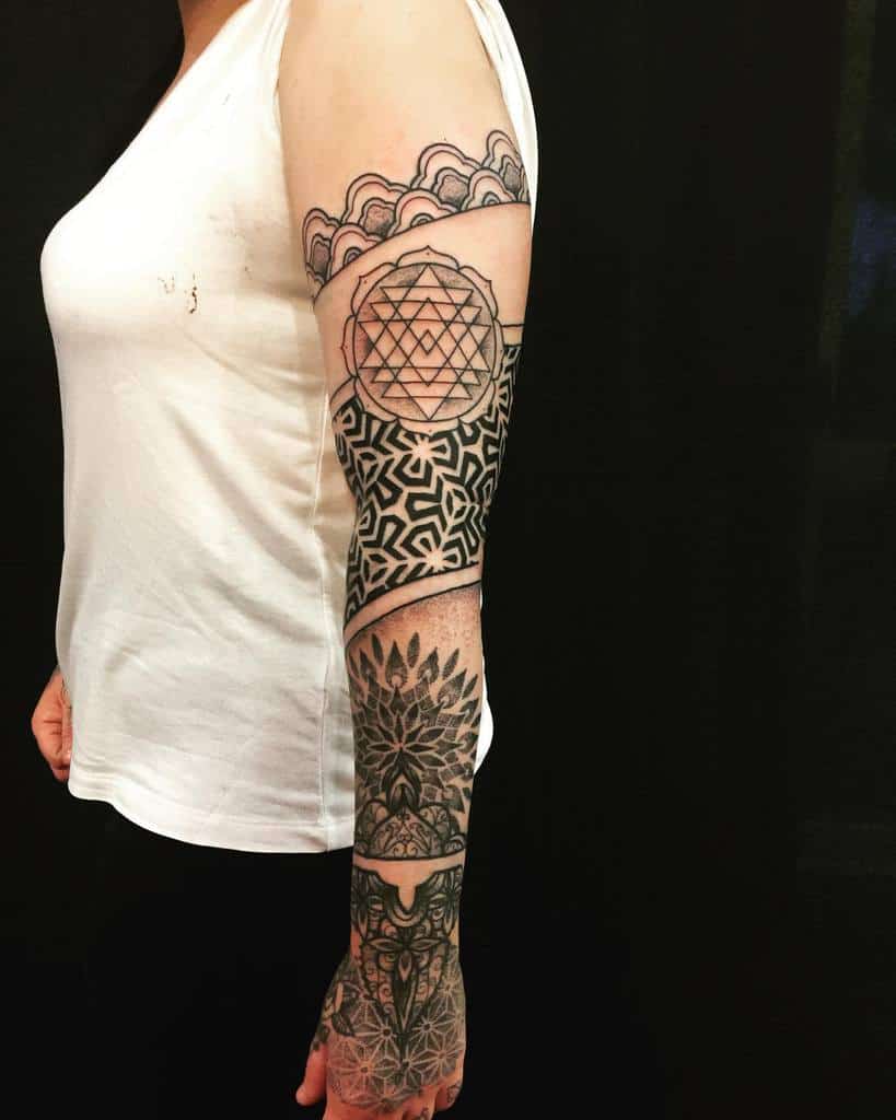 A geometric half sleeve tattoo featuring intricate patterns, including mandala designs, a Sri Yantra symbol, and bold black linework, extending from the upper arm to the wrist
