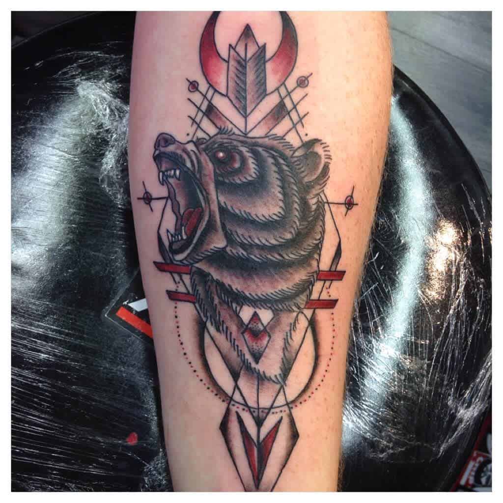 Tattoo of a roaring bear head seamlessly integrated with geometric shapes and bold red accents on an arm, capturing the raw power and spirit of the majestic bear